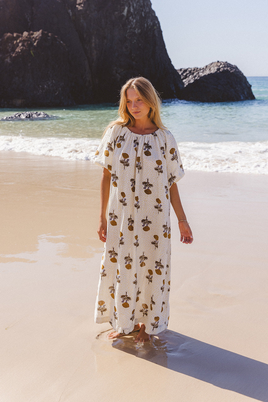 Dahlia Maxi Dress Marigold — full length at beach with sea stacks, on-shoulder styling, white hand block print linen maxi with golden botanical pattern | Daughters of India