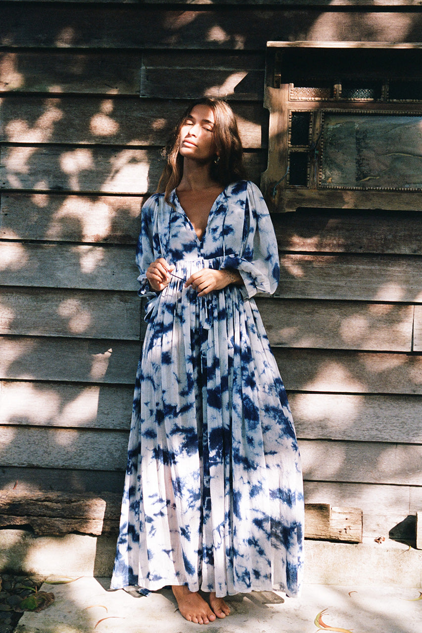 Kyra Maxi Dress Deep Sea — full length front view leaning against rustic wooden wall, deep sea shibori tie-dye cotton maxi dress with flowing tiered skirt, dappled sunlight India | Daughters of India