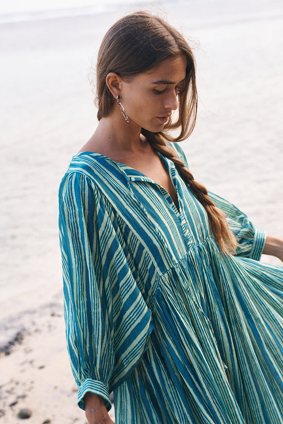 Woman with a braided hairstyle at the beach wearing the Daughters of India Kyra Midi Dress in Spearmint, the green striped fabric flowing in the sea breeze
