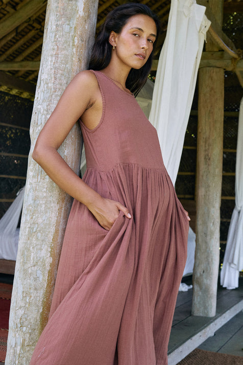 Nadi Dress Chocolate — full length side view leaning against wooden post, chocolate brown double gauze cotton midi dress with hand in pocket, rustic bamboo veranda setting | Daughters of India