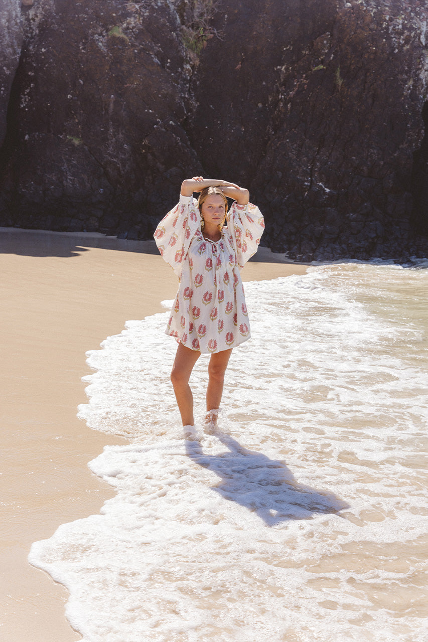 Ria Mini Dress Strawberry Cream — back view beach portrait, model walking along shore wearing strawberry cream floral hand block print cotton mini dress, ocean waves backdrop, artisan Jaipur India | Daughters of India