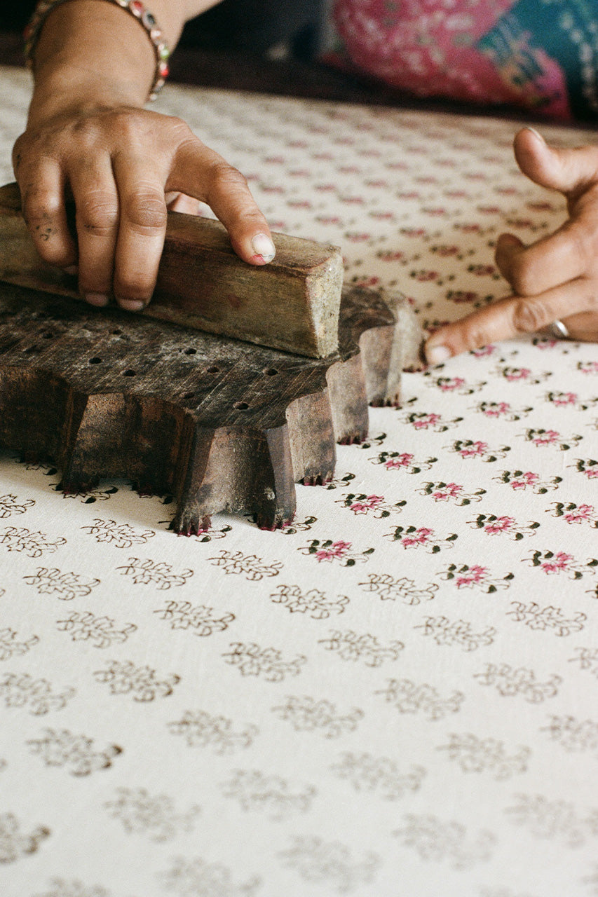 Indian artisan's hands using a carved wooden block to hand-print the pink floral Peony pattern onto cotton fabric, showing the traditional craft behind Daughters of India